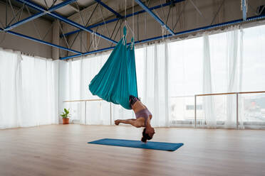 Seitenansicht einer aktiven Frau, die einen Schwanensprung in der Hängematte während eines Aerial-Yoga-Trainings in einem hellen, geräumigen Studio mit Matte übt - ADSF34878