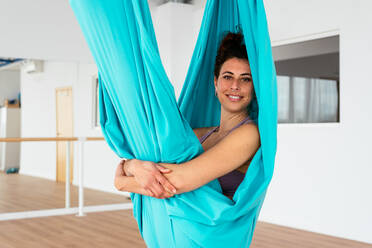 Fröhliche, aktive Frau, die lächelnd in die Kamera schaut, während sie in der Schaukel sitzt, während des Aerial Yoga Trainings in einem hellen, modernen Studio - ADSF34879