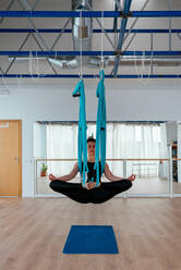 Ganzer Körper einer aktiven Frau in Activewear beim Üben der Antigravitations-Taubenpose auf der Hängematte während des Aerial-Yoga-Trainings im Lichtstudio - ADSF34882