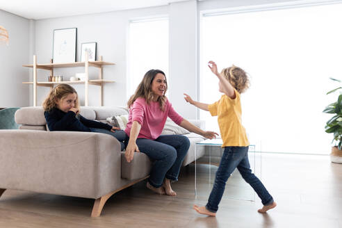 Zärtliche Mutter umarmt niedlichen Sohn, während sie auf einer bequemen Couch mit positiver Tochter sitzt und Zeit im hellen Wohnzimmer verbringt - ADSF34883