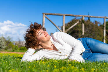 Dreamy happy female in casual clothes lying with eyes closed on green grassy meadow with hand behind head on sunny summer day in nature - ADSF34894