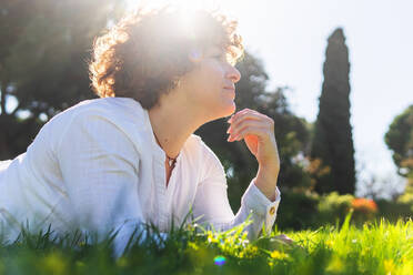 Verträumte, glückliche Frau in Freizeitkleidung, die auf einer grünen Wiese liegt und wegschaut, mit der Hand hinter dem Kopf an einem sonnigen Sommertag in der Natur - ADSF34895