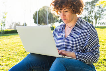 Konzentrierte Frau in Freizeitkleidung, die auf einem Netbook tippt, während sie an einem sonnigen Sommertag auf einer grünen Wiese im Park sitzt - ADSF34898