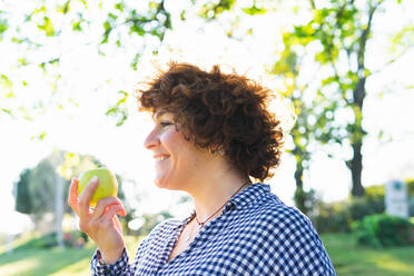 Side view of smiling female with green apple in hand standing on grassy field in park on blurred background on summer day - ADSF34899