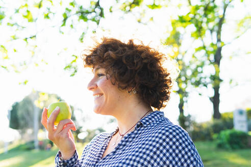 Seitenansicht einer lächelnden Frau mit einem grünen Apfel in der Hand, die auf einer Wiese im Park steht, mit unscharfem Hintergrund an einem Sommertag - ADSF34899