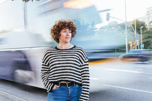Nachdenkliche Frau, die in die Ferne schaut, während sie am Straßenrand in der Nähe eines unscharfen Linienbusses steht, der sich in der Stadt an einem Sommertag bewegt - ADSF34901