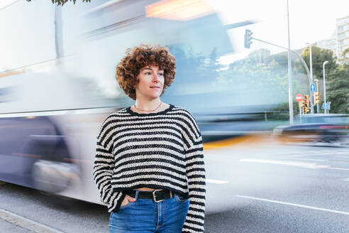 Pensive female looking into distance while standing on roadside near blurred public bus in motion in city on summer day - ADSF34901