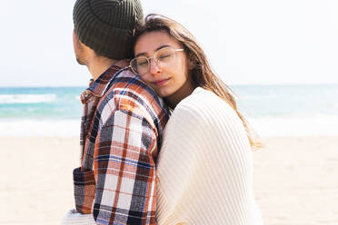 Romantic couple in casual wear cuddling while standing together on sandy beach near waving sea on sunny day in coastal area - ADSF34903