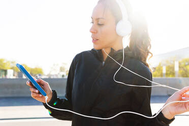 Fit female runner in sportswear browsing smartphone while listening to music via headphones during training on sunny summer day in city - ADSF34905