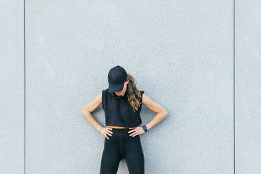 Fit anonymous female athlete in black sportswear and cap standing with hands on waist near gray wall of building during outdoor training in city and looking down - ADSF34909
