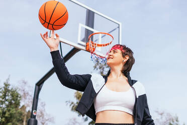 Niedriger Winkel Seitenansicht der weiblichen Spielerin wirft Ball in den Korb beim Basketball spielen gegen bewölkten Himmel und grüne Bäume - ADSF34912