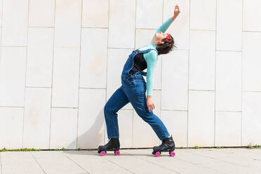 Frau mit kurzen Haaren in Jeans-Overall hebt den Arm, während sie auf vier Rollen auf dem Bürgersteig gegen eine Hauswand tanzt - ADSF34913