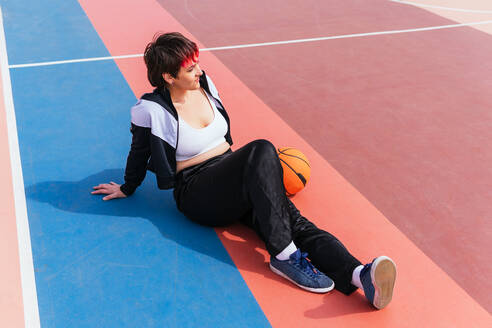 Dame in Sportkleidung mit kurzen gefärbten Haaren, die auf einem Sportplatz mit einem Basketball liegt und wegschaut, von oben - ADSF34914