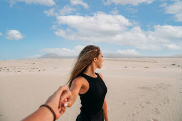 Junge weibliche Touristin, die die Berge und Dünen von Corralejo betrachtet, während sie unter bewölktem Himmel bei Sonnenuntergang auf den Kanarischen Inseln Fuerteventura einen nicht erkennbaren Geliebten in der Hand hält - ADSF34934
