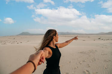 Junge weibliche Touristin, die auf die Berge und Dünen von Corralejo zeigt, während sie unter bewölktem Himmel bei Sonnenuntergang auf den Kanarischen Inseln Fuerteventura einen nicht erkennbaren Geliebten in der Hand hält - ADSF34935