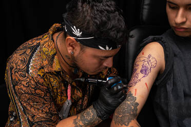 Concentrated Peruvian creative master making tattoo with professional machine on arm of shirtless male client during appointment in modern salon - ADSF34940