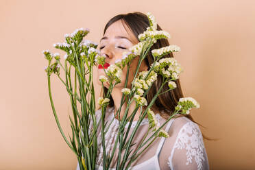 Charmante Braut in weißem Kleid riecht Bouquet von frischen Blumen, während mit geschlossenen Augen auf beige Hintergrund während der Hochzeitsfeier im Studio stehen - ADSF34944