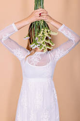 Charmante unerkennbar Braut in eleganten weißen Hochzeitskleid bedeckt Augen mit Strauß frischer Blumen auf beige Hintergrund in hellen Studio - ADSF34945