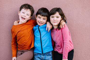 Positive children in casual wear hugging and looking at camera with smile while standing on pink background on street of city - ADSF34947