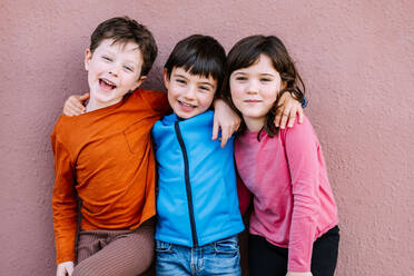 Positive Kinder in Freizeitkleidung, die sich umarmen und lächelnd in die Kamera schauen, während sie auf einem rosa Hintergrund auf einer Straße in der Stadt stehen - ADSF34947