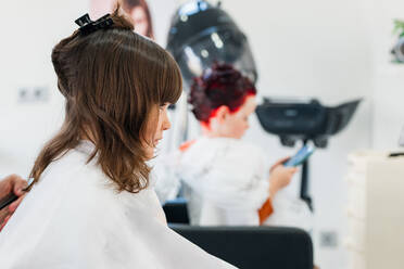 Hands of crop anonymous hairdresser cutting dark hair of cute girl wrapped in white cape sitting in light modern hairdressing salon - ADSF34949