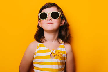 Nettes ernstes Mädchen mit dunklem Haar in gestreifter Kleidung und trendiger Sonnenbrille auf gelbem Hintergrund in modernem Lichtstudio stehend - ADSF34954