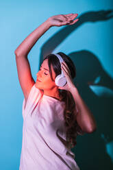 Attraktive Frau hört Musik mit drahtlosen Kopfhörern auf blauem Hintergrund im Studio mit roten Lichtern - ADSF34968