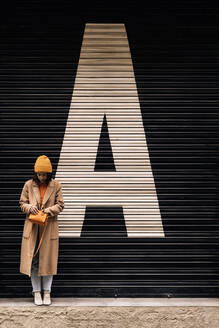 Full body of female with handbag in coat and hat standing near building with closed metal shutter on street of city - ADSF34977