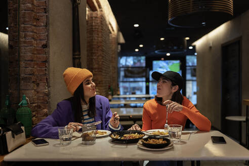 Happy female friends in headwear looking at each other while eating delicious dishes at table with smartphones in modern light cafe - ADSF34983