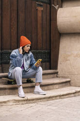 Ganzer Körper einer Frau in Freizeitkleidung und Hut Textnachrichten auf dem Smartphone, während sie auf einem Skateboard in der Nähe eines Gebäudes in der Stadt sitzt - ADSF34985