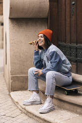 Full body of cheerful female in casual wear and hat listening recording audio message on smartphone while sitting on skateboard near building - ADSF34986