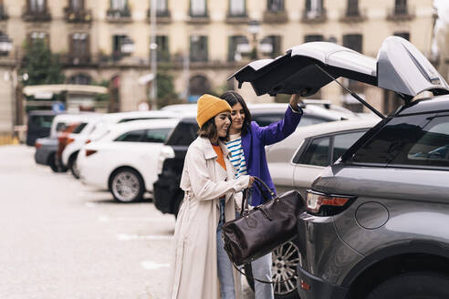 Positive Freundinnen in stilvoller Kleidung, die eine Tasche in den Lastwagen eines modernen Autos packen, das auf der Straße einer Stadt mit einem Gebäude geparkt ist - ADSF34989