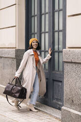 Ganzkörperpositive Frau mit Tasche in stilvollem Mantel und Hut in der Nähe der schwarzen Eingangstür eines Gebäudes in der Stadt stehend - ADSF34990