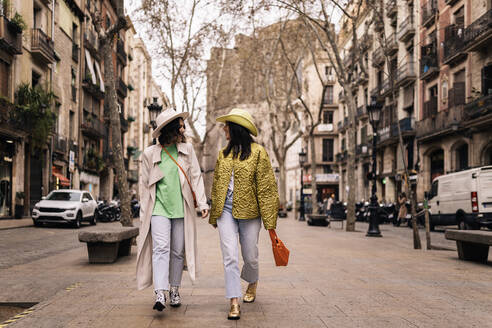 Full body of female friends in stylish hats looking at each other while strolling along buildings on paved walkway in city - ADSF34991