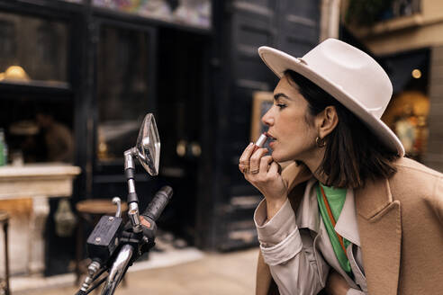 Seitenansicht einer eleganten Frau mit Hut und Mantel, die sich die Lippen bemalt, während sie in den Spiegel eines auf der Straße geparkten Fahrrads schaut - ADSF34993