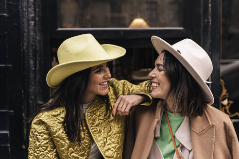 Smiling female friends in trendy hats and outerwear looking at each other while standing together near entrance of building in city - ADSF34997