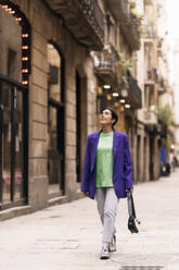 Fröhliche Frau mit Handtasche in stilvollem Mantel, die lächelnd nach oben schaut, während sie auf der Straße der Stadt spazieren geht - ADSF34998