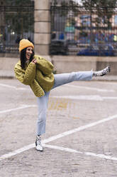 Full body of positive active female in outerwear and hat doing leg kick while standing on paved sidewalk in city - ADSF35004