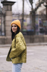 Seitenansicht einer Frau in modischem Mantel und Mütze, die auf der Straße einer Stadt spazieren geht und in die Kamera schaut - ADSF35006