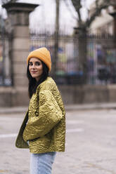 Side view of female in stylish coat and beanie walking on the street of city looking at camera - ADSF35006