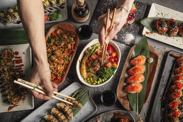 Von oben der Ernte anonyme Menschen essen verschiedene orientalische Lebensmittel mit Stäbchen am Tisch mit Sushi und Poke in der Nähe von Udon und Jiaozi im Restaurant - ADSF35009