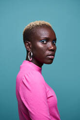 Emotionless African female in pink wear with short dyed hair looking at camera while standing on blue background in studio - ADSF35015