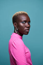 Emotionslose afrikanische Frau in rosa Kleidung mit kurzen gefärbten Haaren, die in die Kamera schaut, während sie auf blauem Hintergrund im Studio steht - ADSF35015