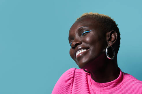 Positive afrikanische Frau mit hellem Make-up und kurzen gefärbten Haaren, die mit geschlossenen Augen nach oben schaut, während sie auf rotem Hintergrund in einem hellen Studio steht - ADSF35016