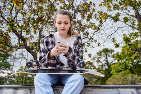 Junge Frau mit schwarzem Skateboard surft auf dem Smartphone, während sie in einem Skatepark sitzt - ADSF35025