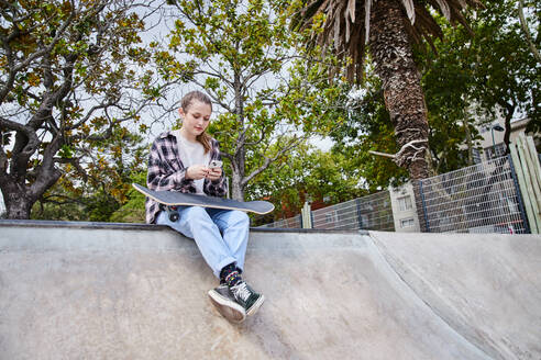 Ganzkörperdarstellung einer jungen Frau mit schwarzem Skateboard, die auf ihrem Smartphone surft, während sie in einem Skatepark sitzt - ADSF35026