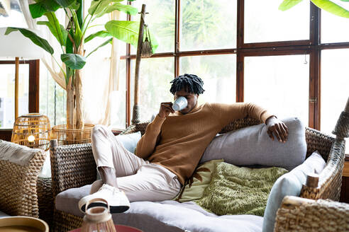 Junger Afroamerikaner, der frischen Kaffee trinkt, während er auf einem bequemen Sofa in einem hellen Wohnzimmer mit Topfpflanze zu Hause sitzt - ADSF35037