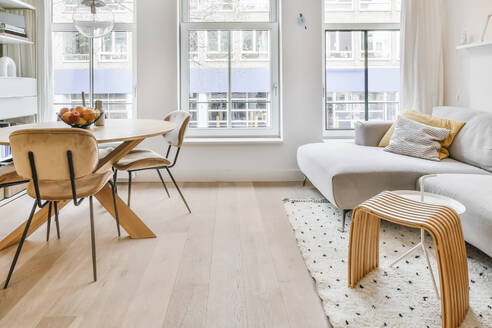 Holzstühle und Tisch vor bequemer Couch auf Teppich an Wand mit dekorativen Rahmen in hellem geräumigen Wohnzimmer platziert - ADSF35055