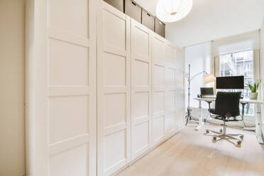 Laptop and computer placed on table near window in light stylish home office with closed white closet in modern apartment - ADSF35058