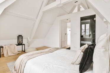 Bequemes Bett mit weißer Decke und Kissen in hellem geräumigen Schlafzimmer mit geöffneter Tür und Bank mit Dekoration in der Wohnung platziert - ADSF35066